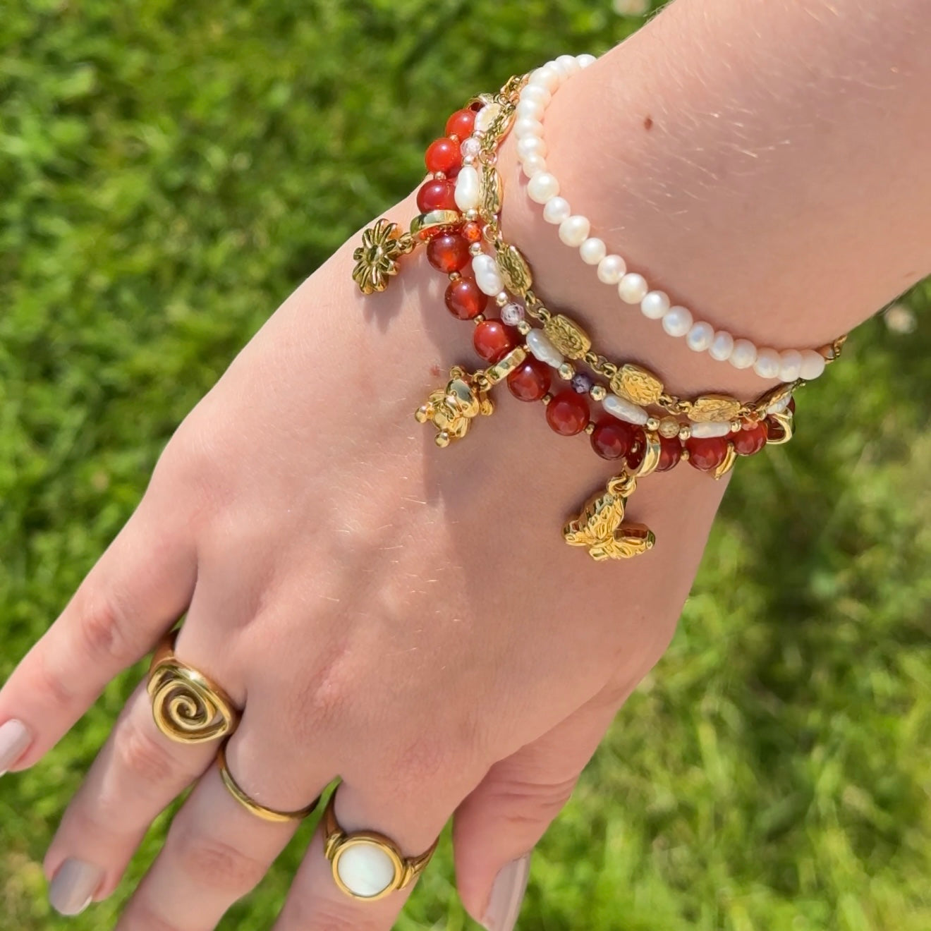 Hand wearing multiple bracelets with red beads and gold accents against a grassy background.