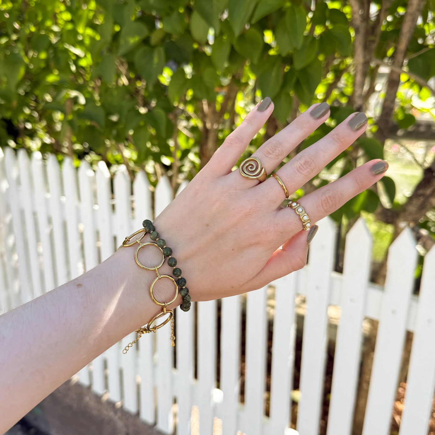 Hand wearing gold bracelets and rings with a white picket fence and greenery in the background
