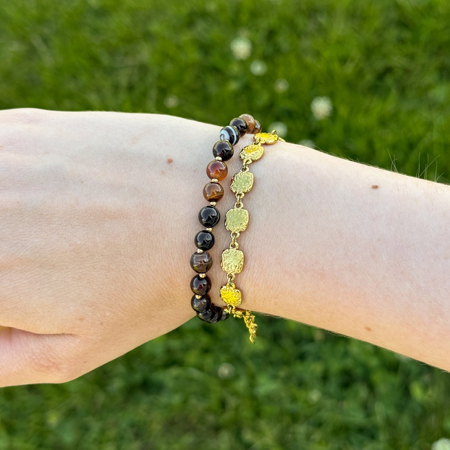 Two bracelets on a wrist against a grassy background. One brown beaded bracelet and one gold vintage-looking medallion bracelet.