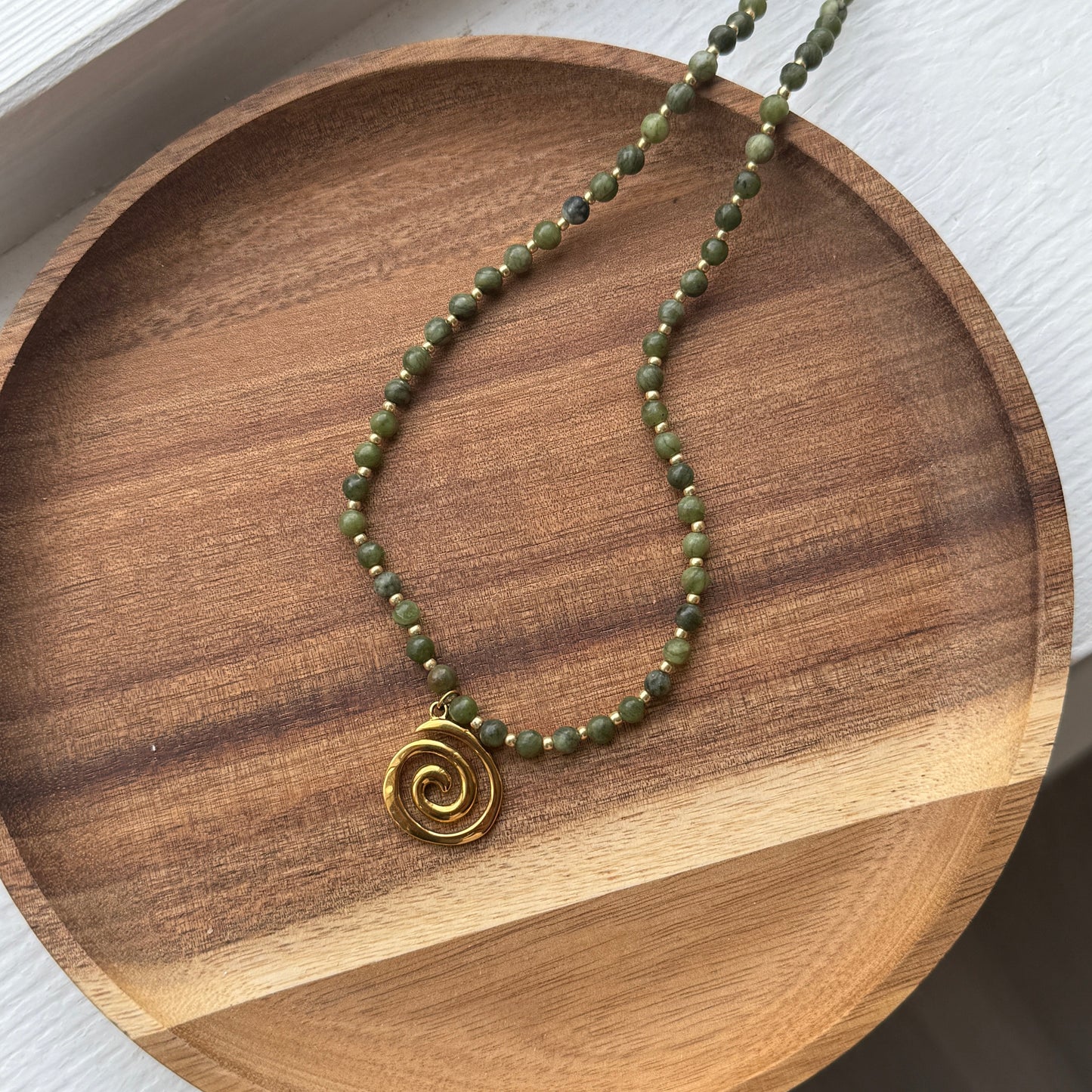 A green beaded necklace with a gold-colored spiral pendant, displayed on a wooden round tray.