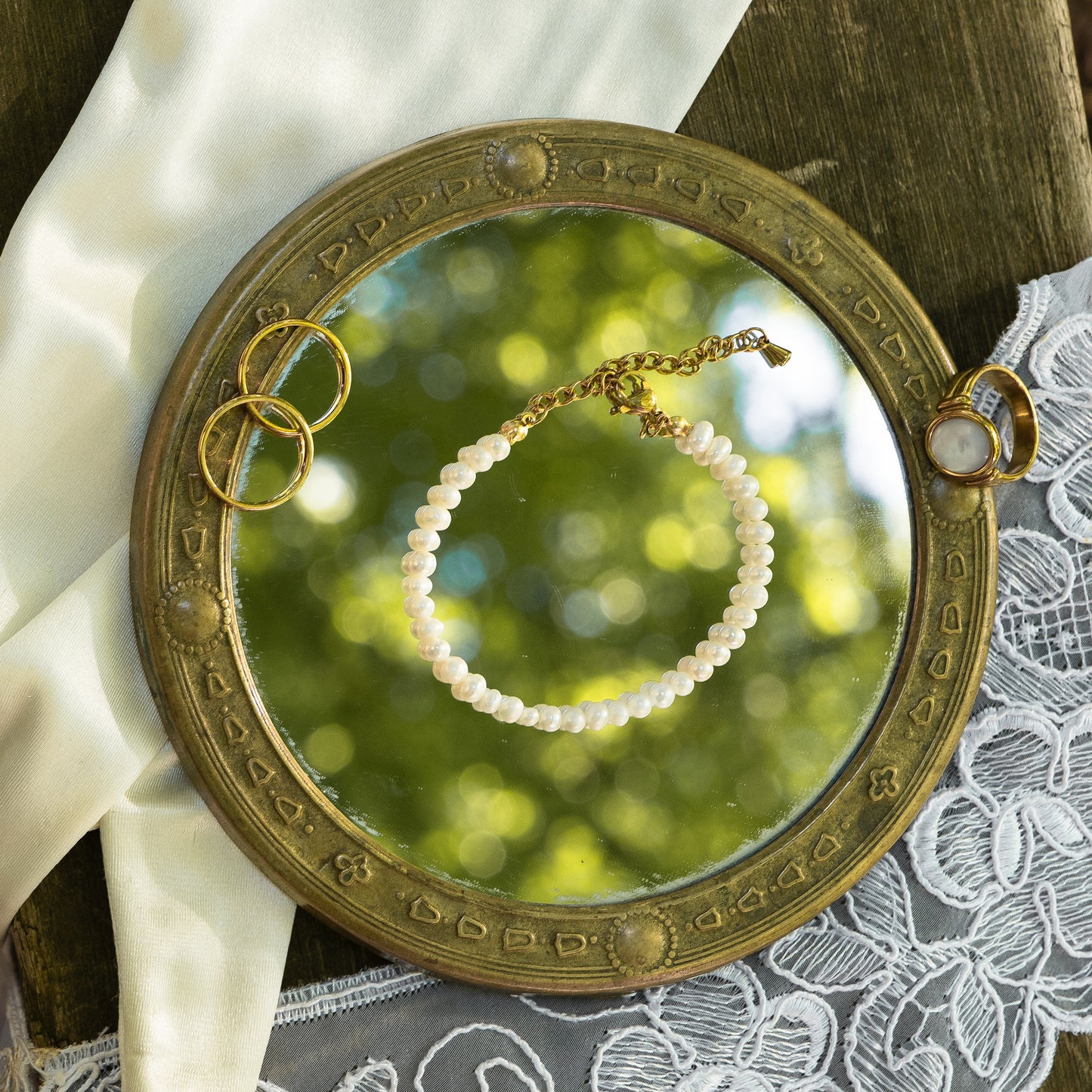 Round mirror with decorative frame on a lace fabric, reflecting a freshwater pearl bracelet and rings.