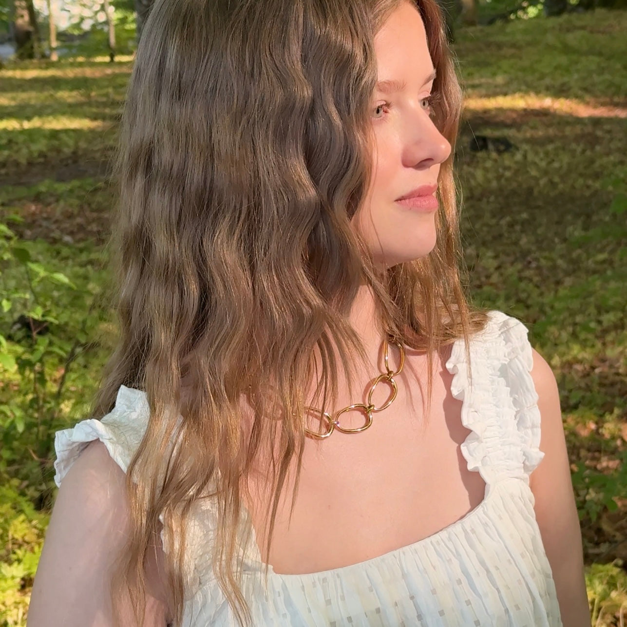 Woman wearing a white dress with ruffled sleeves and a gold link necklace in a park setting