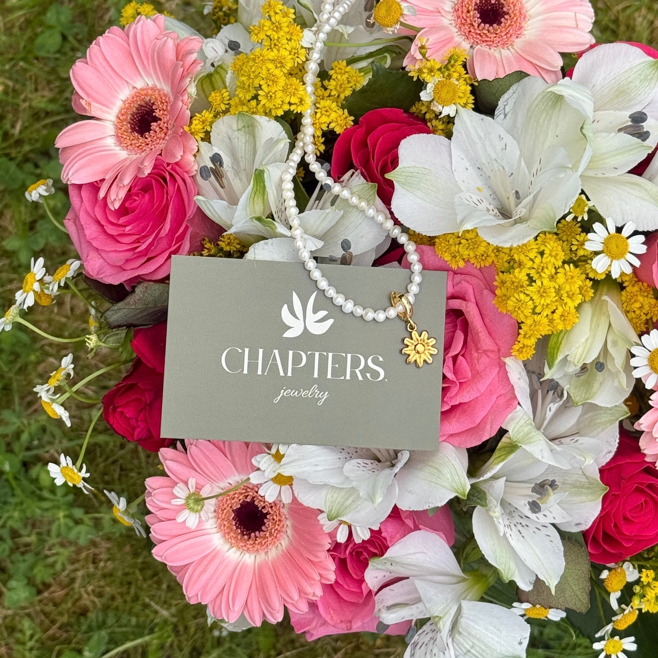Floral arrangement with a 'Chapters Jewelry' card and freshwater pearl necklace with a flower pendant on top.