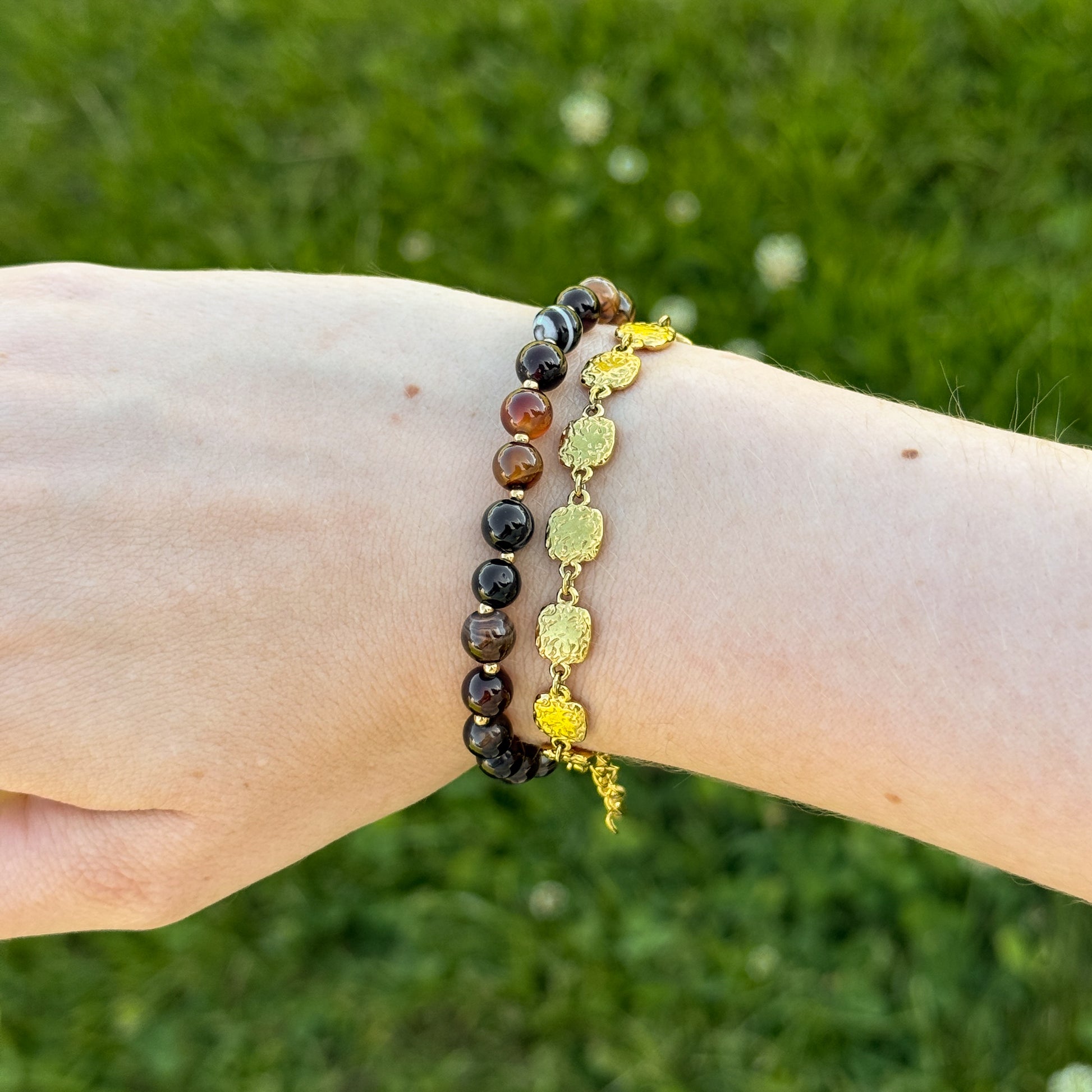 Two bracelets on a wrist against a grassy background. One brown beaded bracelet and one gold vintage-looking medallion bracelet. 
