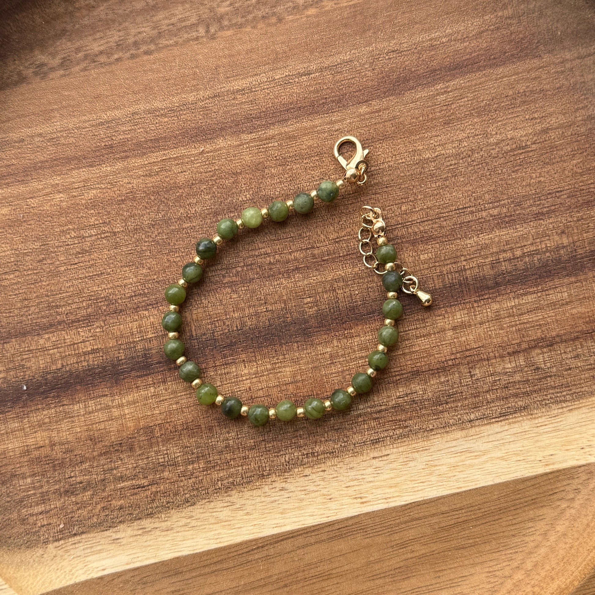 A green beaded bracelet with gold-colored accents and a gold clasp, displayed on a wooden tray.