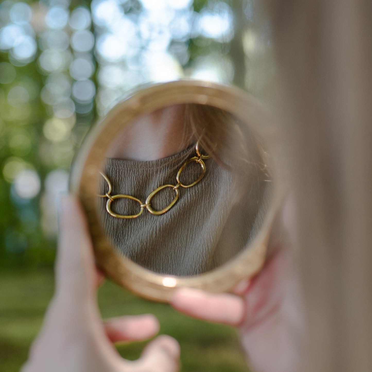 Person holding a round mirror reflecting a gold chain necklace against a blurred natural background