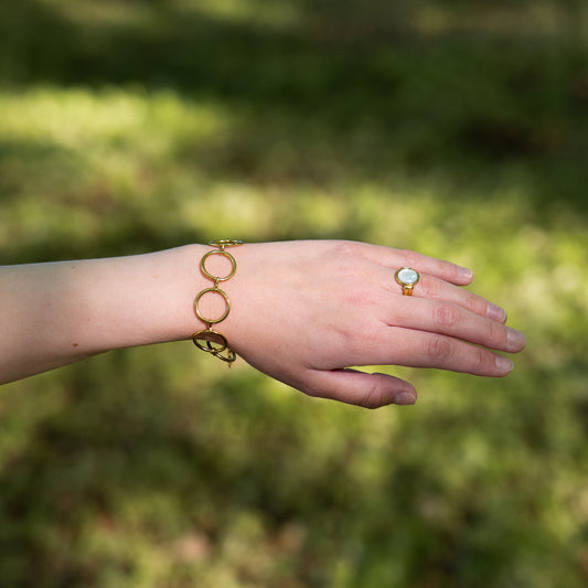 Hand wearing a gold bracelet with large circle chains, and ring with a blurred green background