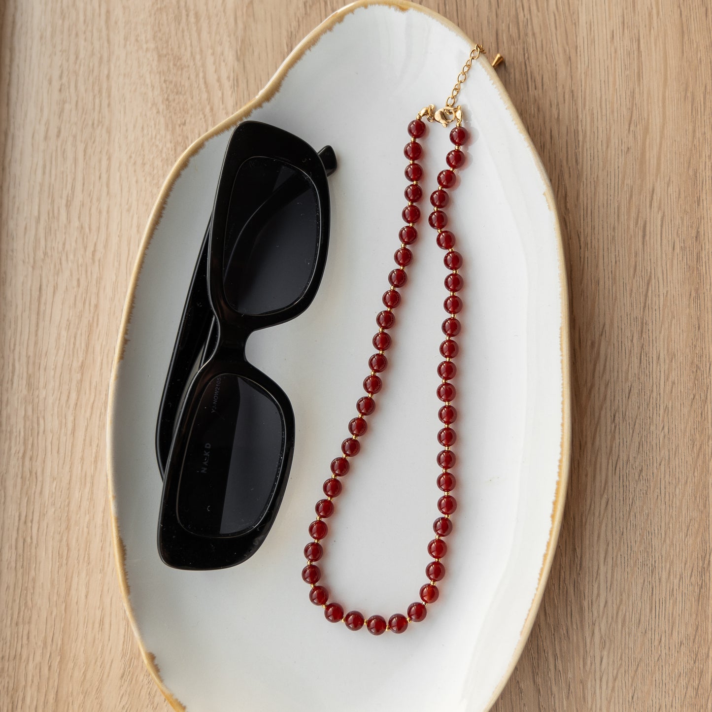 Black sunglasses and a red beaded necklace on a white dish with gold rim.