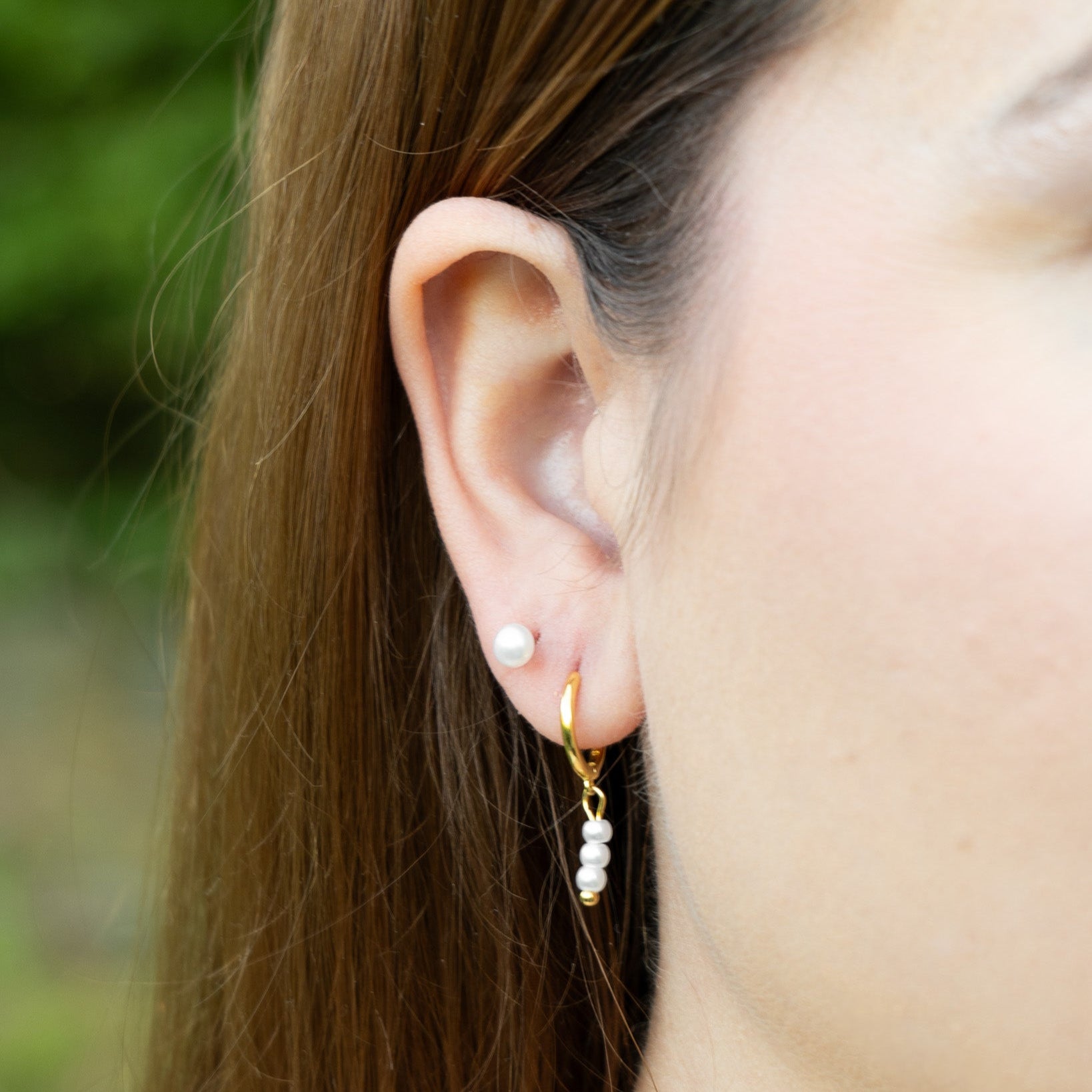 Close-up of an ear wearing a gold earring with pearl drops against a blurred natural background
