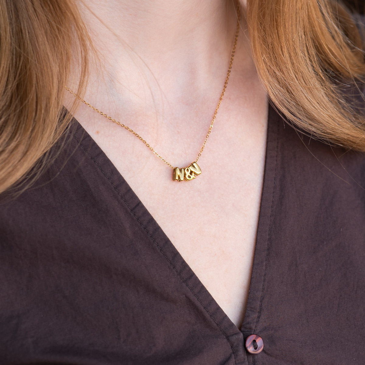 A close-up of a person wearing a delicate gold necklace with a small initial pendant that reads 'N&A', paired with a dark brown button-up shirt.
